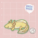 A golden yellow axolotl sticker - shown on a pink measuring grid background with a quarter for scale. Art by Byte Size Treasure
