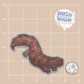 A brown salamander sticker - shown on a tan measuring grid background with a quarter for scale. Art by Byte Size Treasure