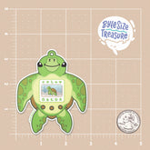 A silly sticker featuring tamagotchi in the shape of a sea turtle - shown on a tan measuring grid background with a quarter for scale. Art by Byte Size Treasure