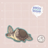 A sticker of a pig-nosed turtle - shown on a pink measuring grid background with a quarter for scale. Art by Byte Size Treasure