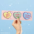 Hand holding a pink bumper sticker with heart-shaped messages &